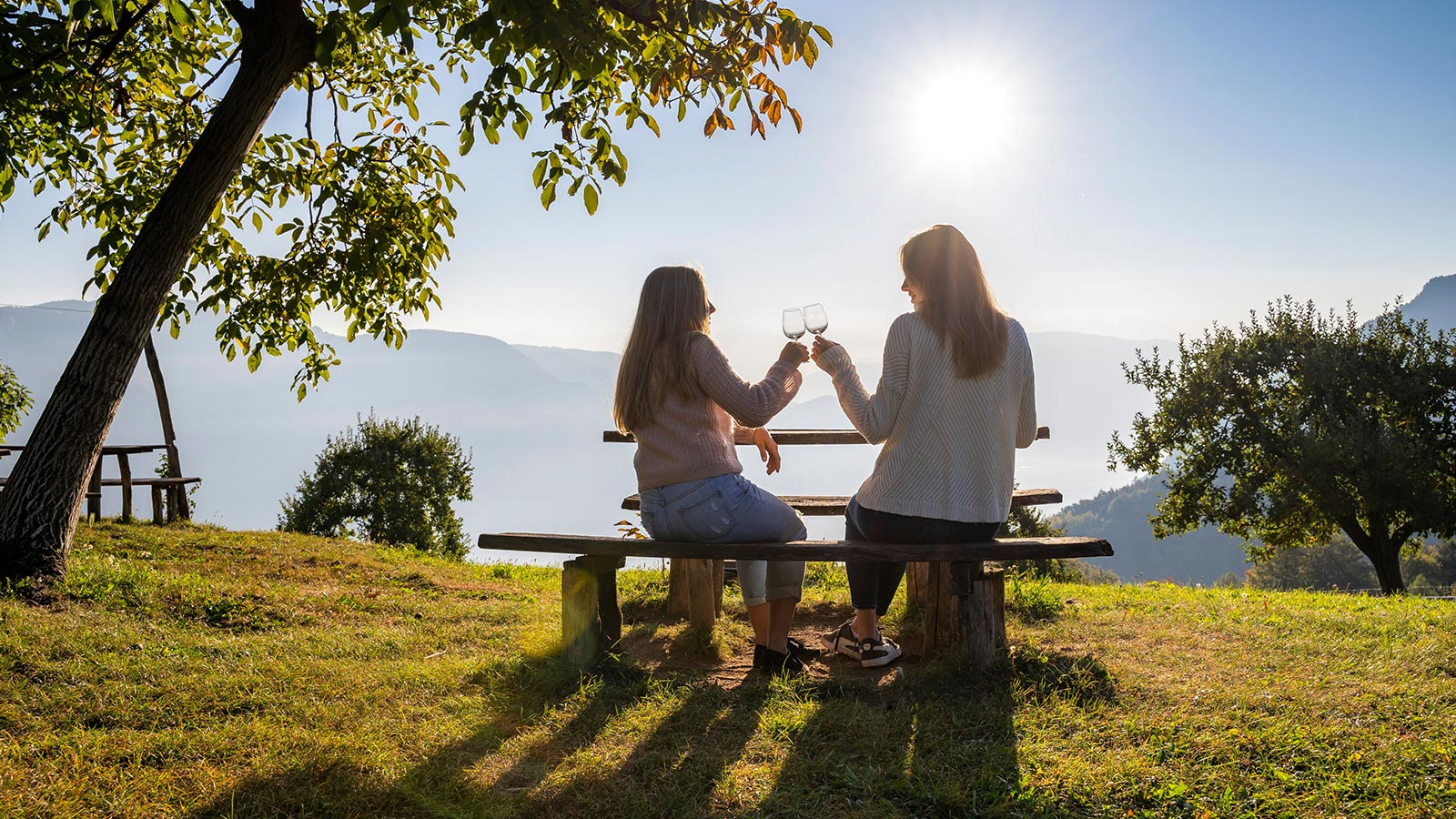 Zwei Mädchen stoßen mit zwei Gläsern Wein an und bewundern den Sonnenuntergang um Tiesens herum